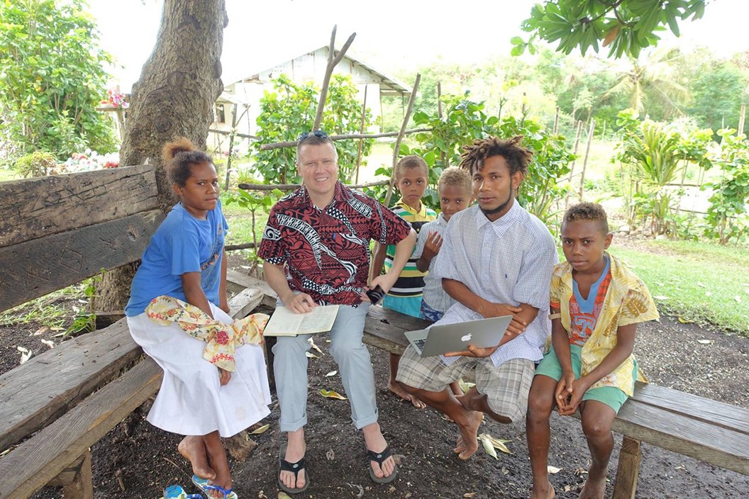 这名男子坐在外面的长凳上，周围是五个瓦努阿图当地人